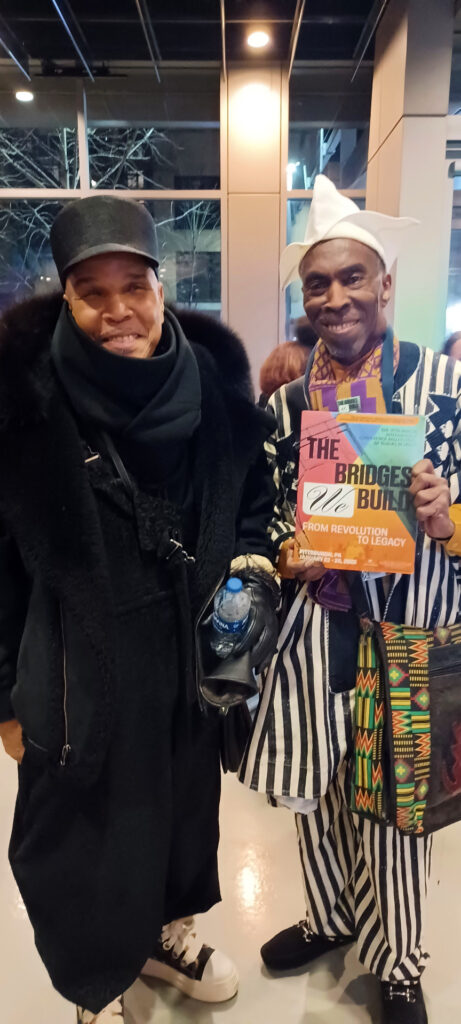 Two smiling individuals stand indoors. The person on the left wears a black cap, coat, and scarf, holding a water bottle. The person on the right has a white hat, colorful patterned attire, and holds a book titled "The Bridges of B.B. Hill: From Revolution To Literacy.