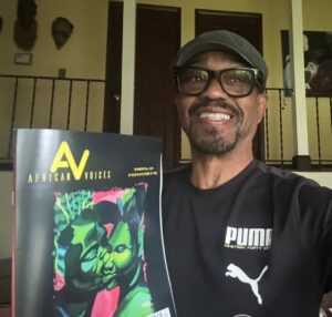 A smiling man in glasses and a cap, Suffolk County’s first African-American poet laureate, holds an “African Voices” magazine with artwork of two faces. He wears a black Puma shirt in a warmly lit room adorned with wall art and masks.