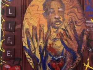 An oval painting shows a dark-skinned figure with closed eyes and raised hands in warm tones. Scrabble tiles spelling “LIFE” run vertically on the left. The background features hearts and swirling patterns in red, blue, and yellow hues.