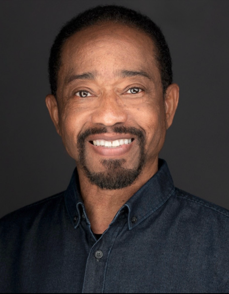 Smiling man with short black hair and a goatee, wearing a dark denim button-up shirt. He has brown skin and is posed in front of a plain dark background—Suffolk County’s first African-American poet laureate, looking warmly at the camera.